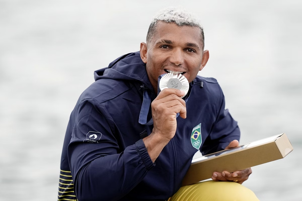 Ajoelhado e sorrindo, Isaquias Queiroz, homem negro de cabelos loiros, posa segurando medalha olímpica de prata